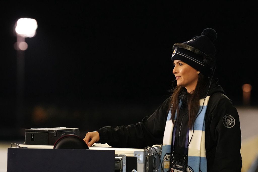 Jamie Anne photographed side on while standing at the decks, with a dark night-time background and one hand extended forward to the mixing desk. She's dressed in a black woolly hat and jacket with a light blue and white Manchester City scarf